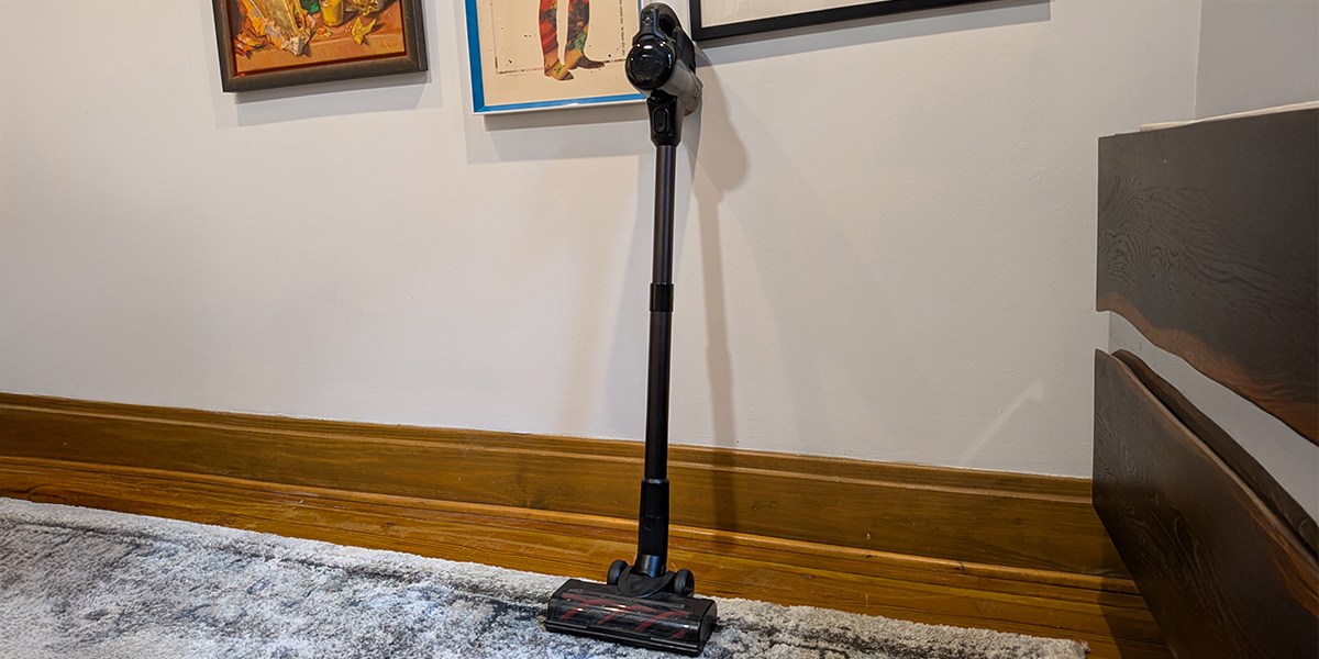 A stick vacuum leaning up against a white wall atop of gray carpet next to a credenza